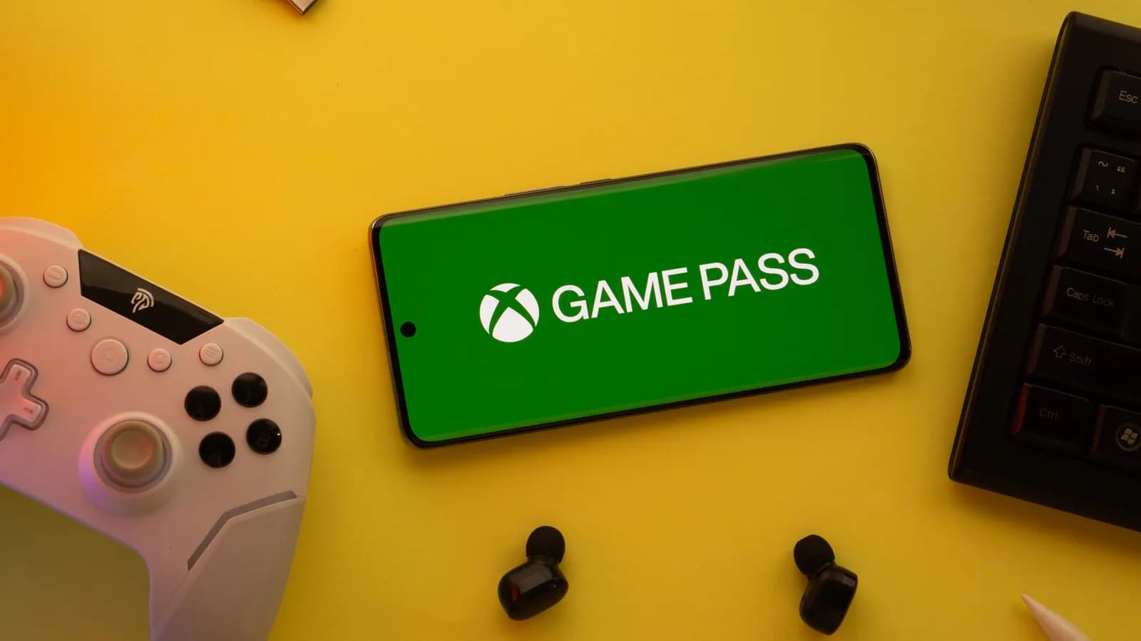 Phone showing the Xbox Game Pass logo on a yellow background, beside a game controller, keyboard and wireless headphones
