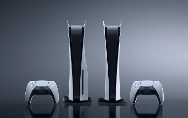 Two PlayStation 5 consoles stand side by side with two white DualSense controllers against a gray background.