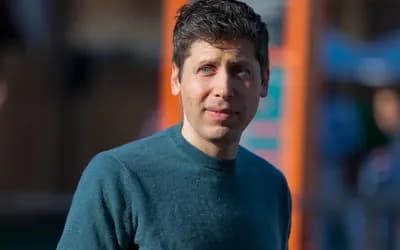 Man with short dark hair in a dark green sweater looks to the side outdoors, with a blurred orange-poled background.