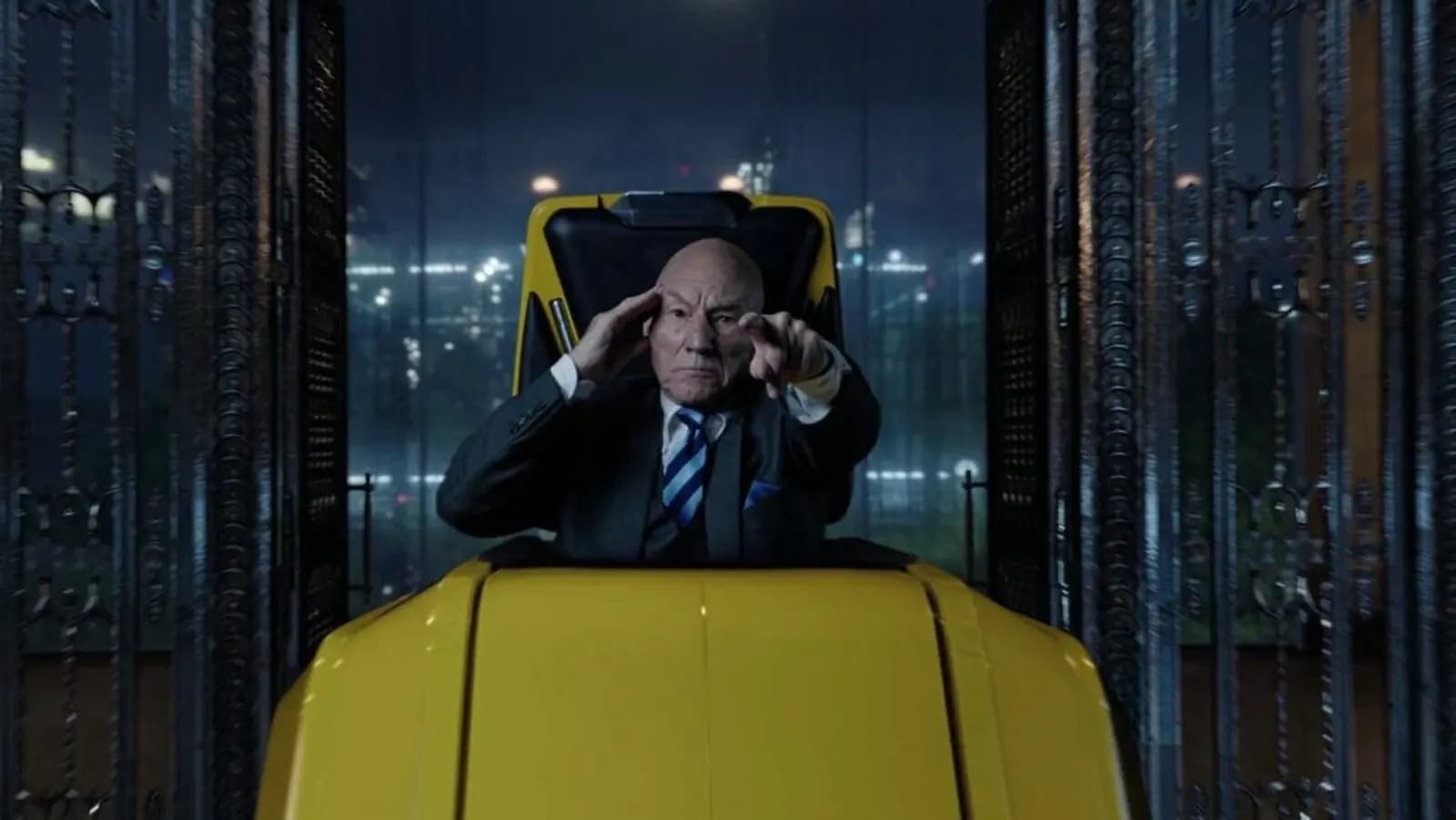 Bald elderly man in a suit sits in a yellow vehicle, one hand at his temple, the other pointing ahead.