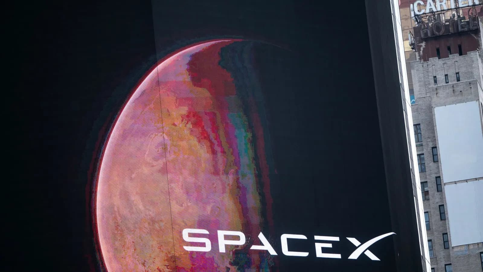 Giant screen on a building shows a red Mars-like planet and the SpaceX logo; an old hotel building stands to the right.