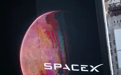 Bir binadaki dev ekranda Mars benzeri kırmızı bir gezegen ve SpaceX logosu görünüyor; sağda eski bir otel binası yer alıyor.