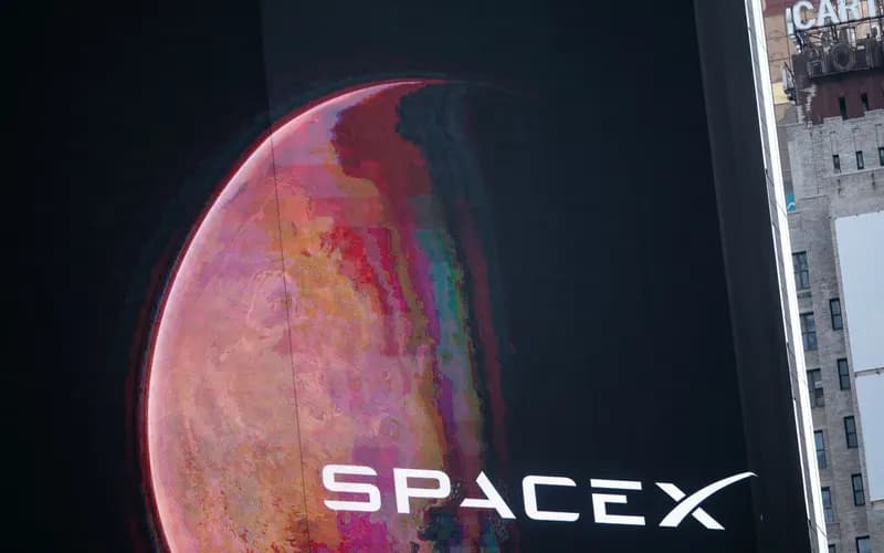 Bir binadaki dev ekranda Mars benzeri kırmızı bir gezegen ve SpaceX logosu görünüyor; sağda eski bir otel binası yer alıyor.