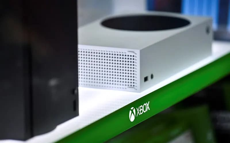 Mağaza rafında yan yana duran beyaz Xbox Series S ve siyah Xbox Series X konsolları, rafın yeşil kenarında Xbox logosu