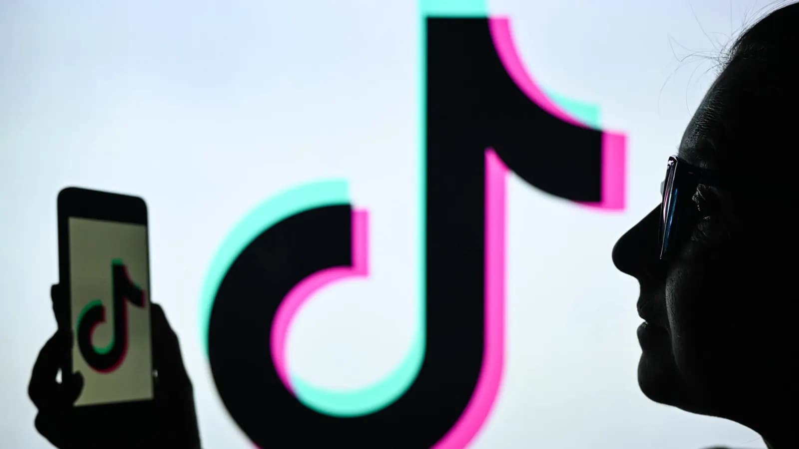 Silhouette of a woman in glasses holding a phone, with the TikTok logo on its screen and a giant screen behind her