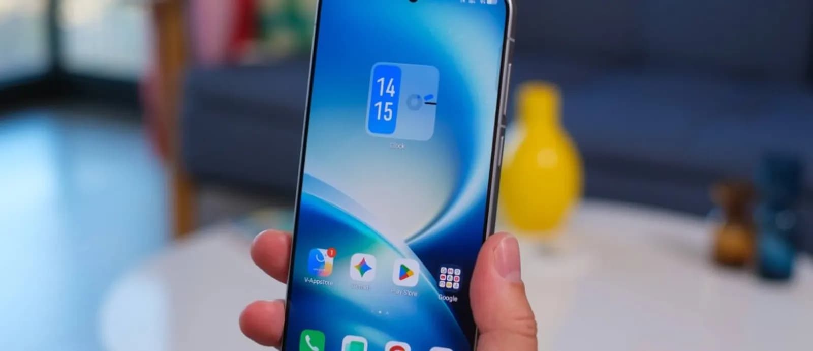 Hand holding a smartphone showing a clock widget and app icons, with a blurred living room in the background