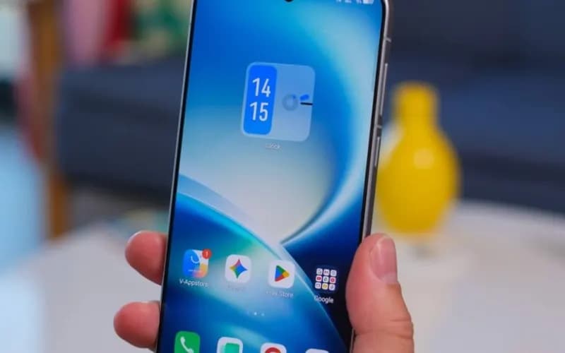 Hand holding a smartphone showing a clock widget and app icons, with a blurred living room in the background