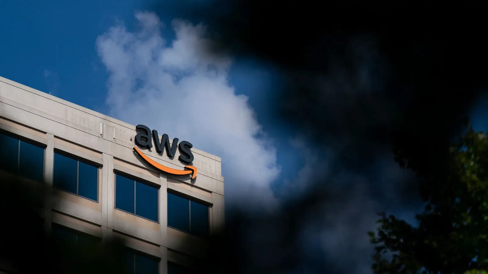 Mavi gökyüzü ve bulutların önünde, çatısında AWS logosu bulunan ofis binası; kenarlarda bulanık yapraklar görünüyor.