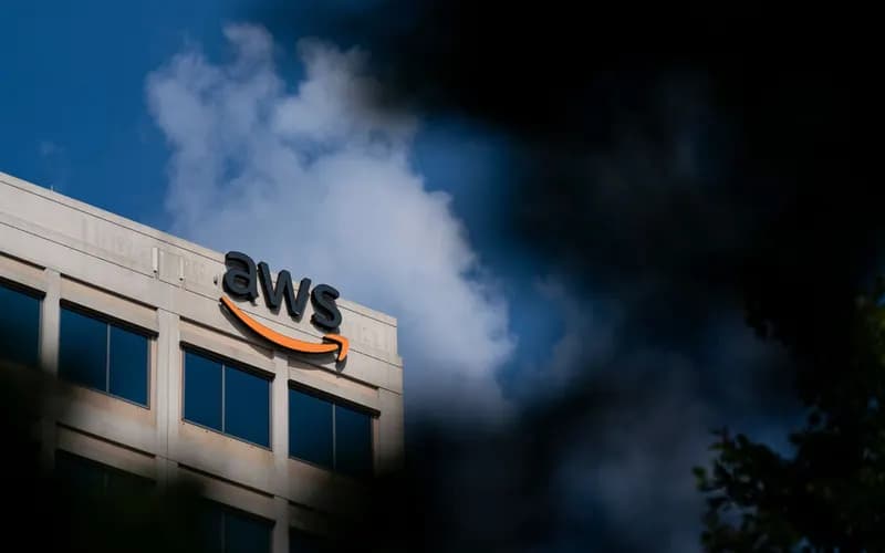 Mavi gökyüzü ve bulutların önünde, çatısında AWS logosu bulunan ofis binası; kenarlarda bulanık yapraklar görünüyor.