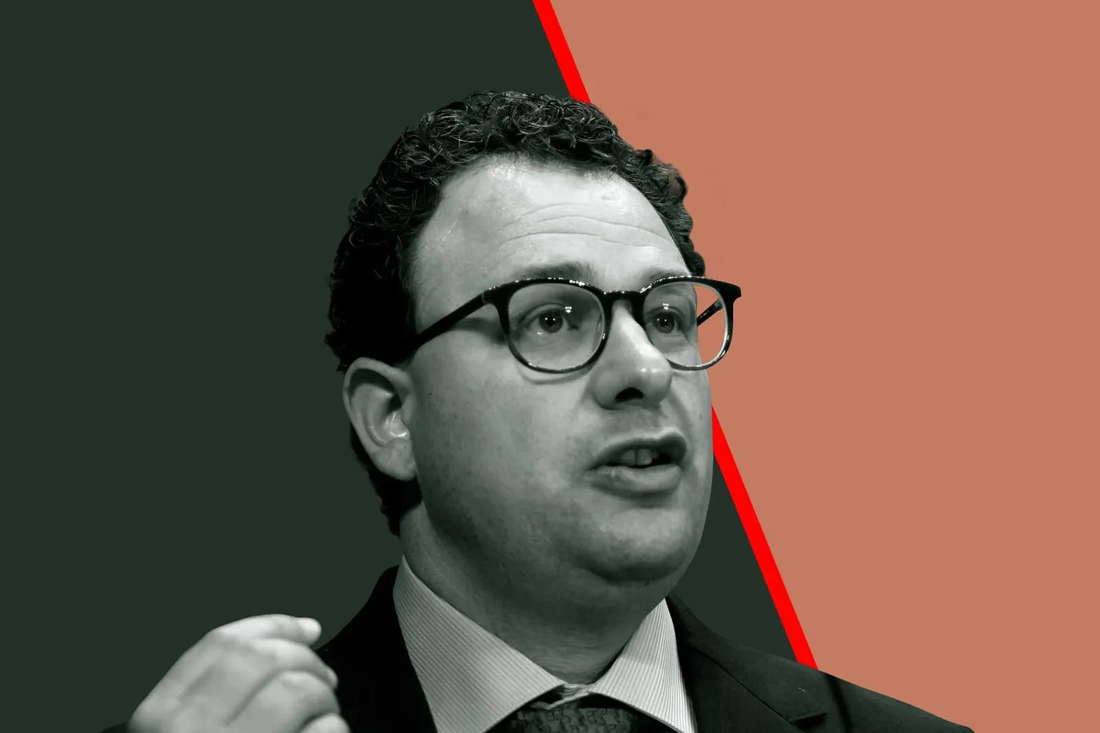 Curly-haired man in glasses and a dark suit raises his hand while speaking against a dark green and orange background.