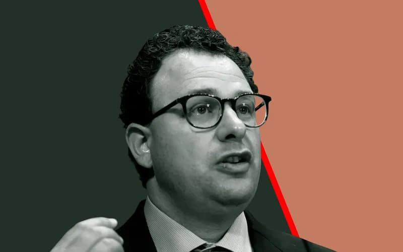 Curly-haired man in glasses and a dark suit raises his hand while speaking against a dark green and orange background.