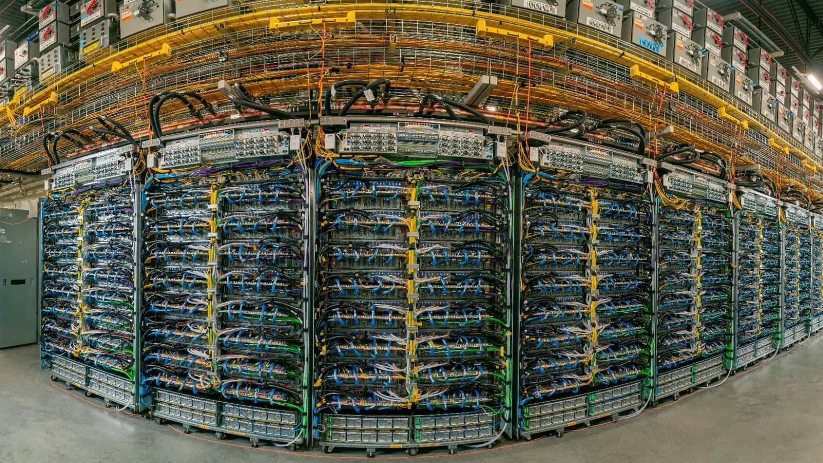 Data center aisle with rows of server racks filled with blue and yellow network cables and cable trays overhead