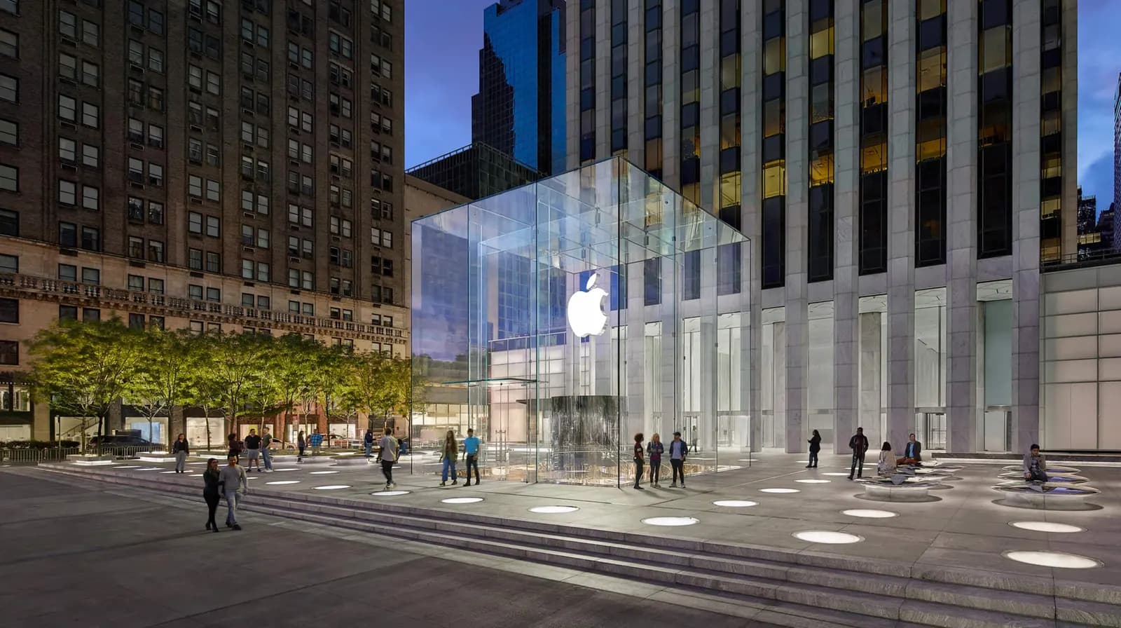 Alacakaranlıkta Manhattan'daki Apple Fifth Avenue mağazasının cam küpü, çevresindeki meydan, basamaklar ve yürüyen insanlar
