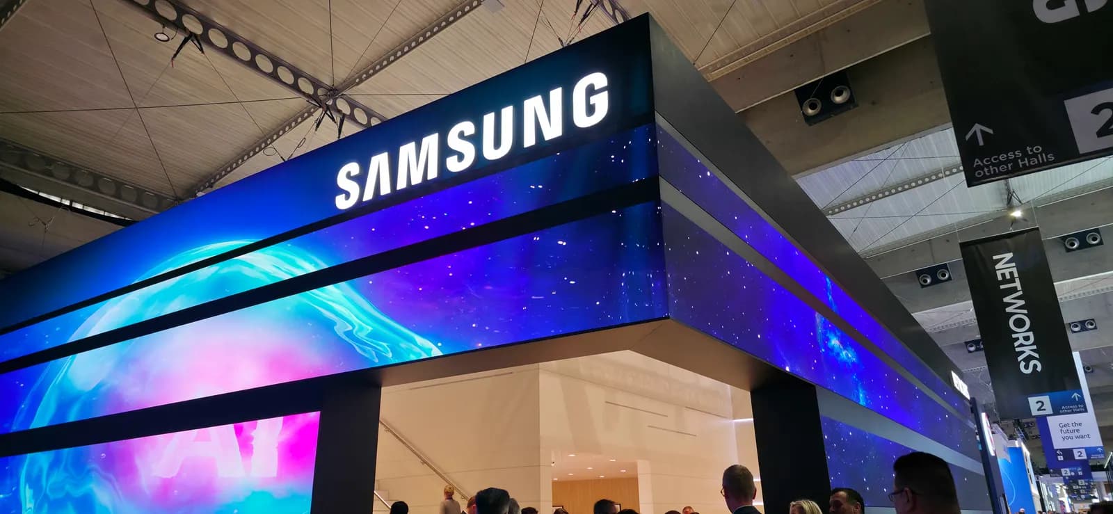 Fuarda Samsung logolu, mavi ve mor LED ekranlarla kaplı büyük stant ile altında dolaşan ziyaretçiler