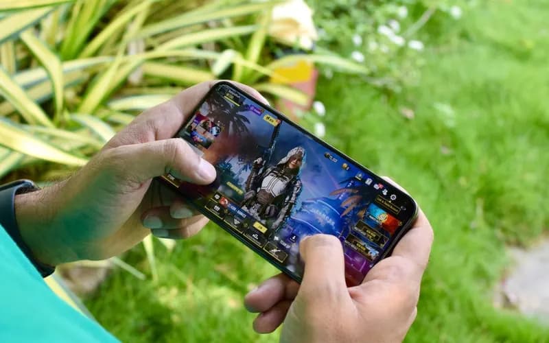 Bahçede çimenlerin yanında iki elle yatay tuttuğu akıllı telefonda mobil oyun oynayan bir kişi