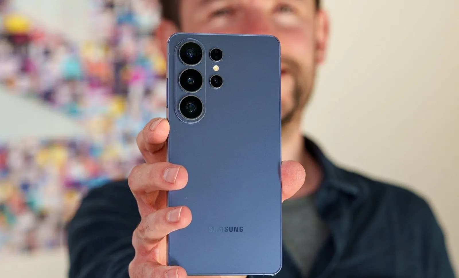Bulanık arka planda bir adam, mavi renkli Samsung Galaxy Ultra telefonun arka yüzünü ve kamera dizisini kameraya doğru tutuyor.