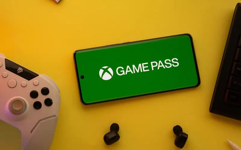 Phone showing the Xbox Game Pass logo on a yellow background, beside a game controller, keyboard and wireless headphones