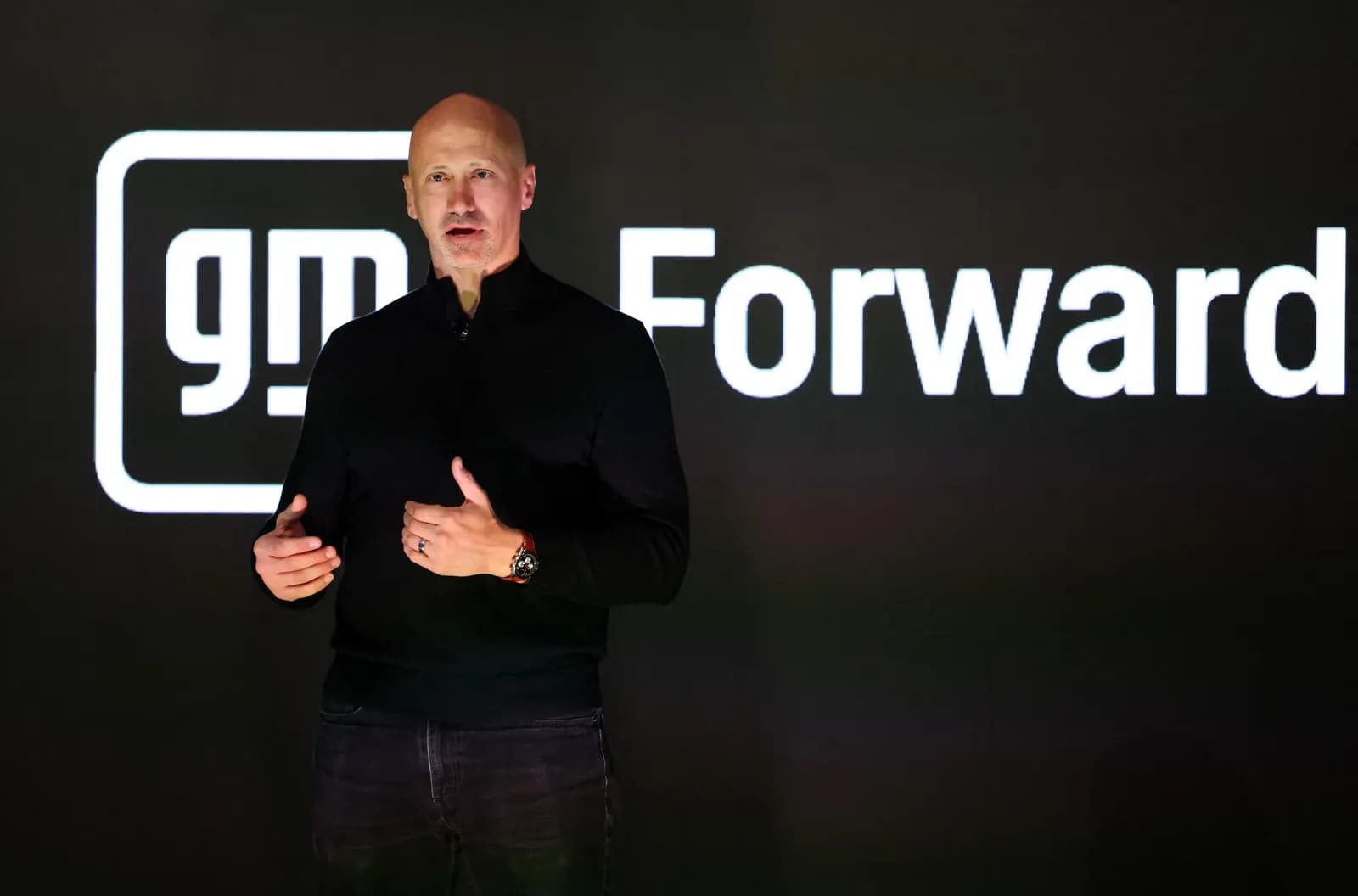 Siyah kazaklı kel bir adam, 'gm Forward' logosunun yer aldığı koyu arka planın önünde konuşma yapıyor.