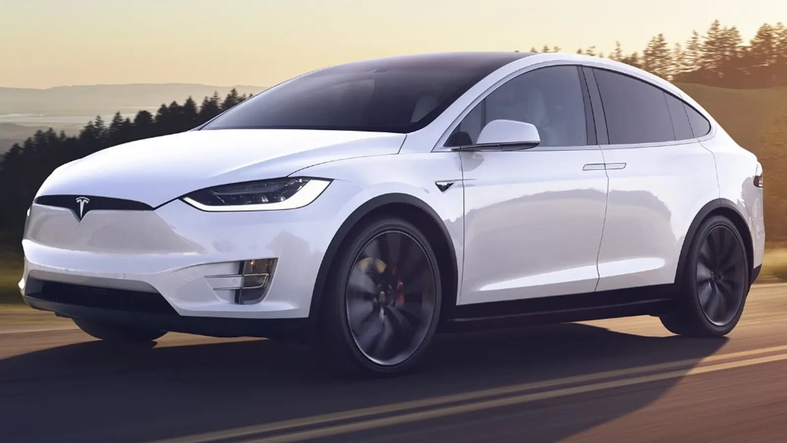 Gün batımında yolda ilerleyen beyaz Tesla Model X elektrikli SUV, arka planda ağaçlar ve tepeler