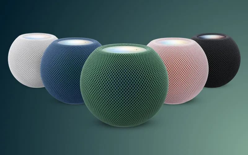 Five small smart speakers in white, blue, green, pink, and black stand side by side on a green background.