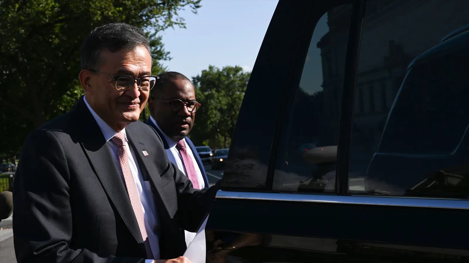 Two men in glasses and dark suits walk beside a black car on a sunny, tree-lined street.