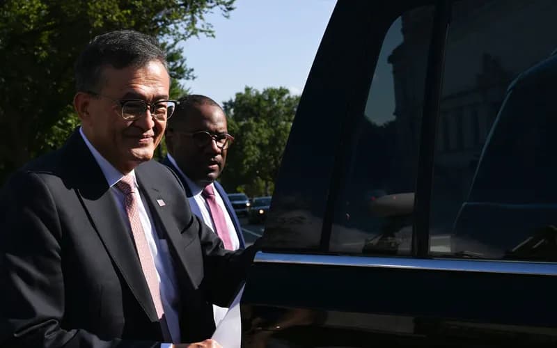 Two men in glasses and dark suits walk beside a black car on a sunny, tree-lined street.