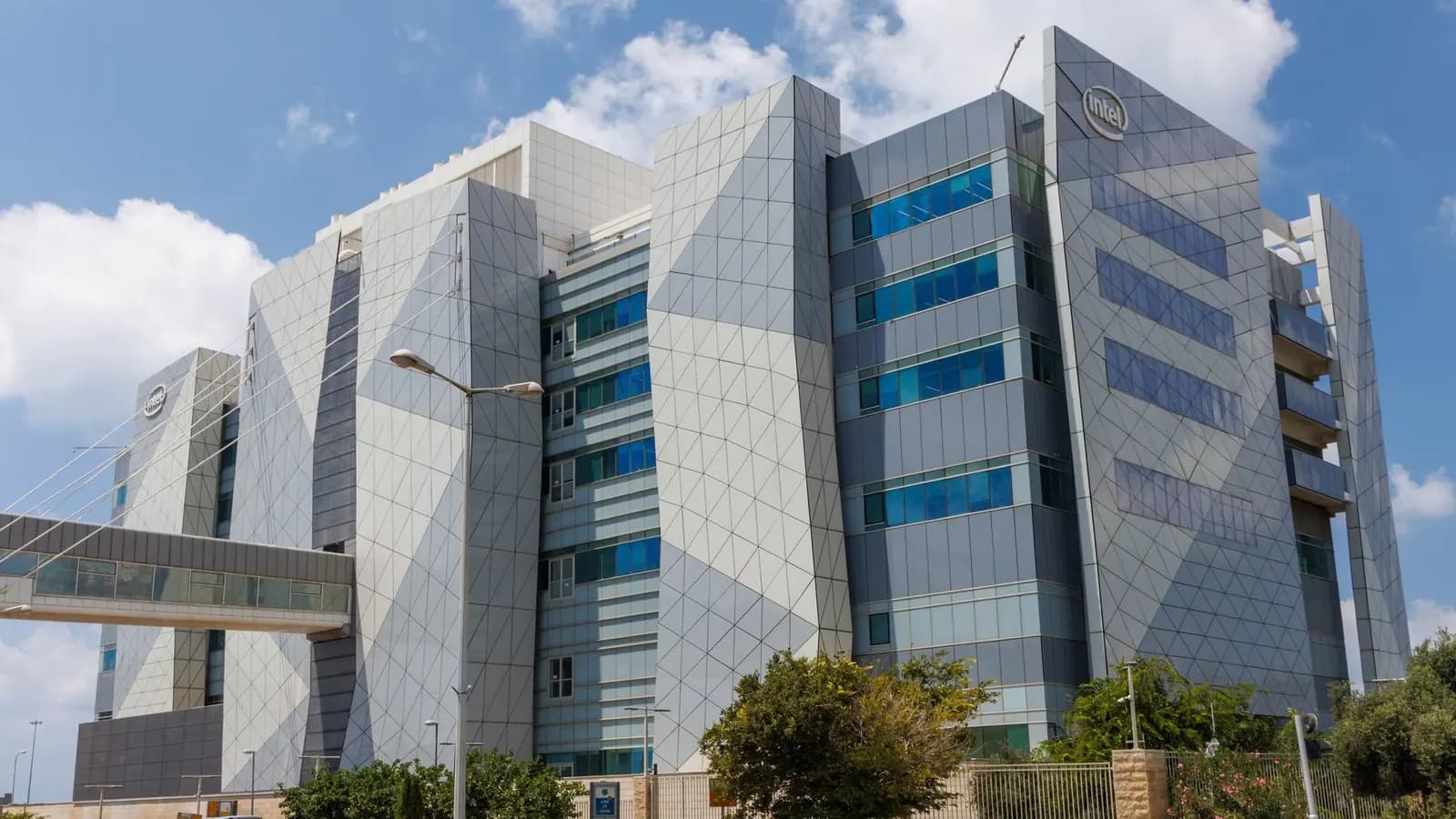 Mavi gökyüzü altında, cephesinde Intel logosu bulunan, gri geometrik kaplamalı modern ofis binası ve cam geçit