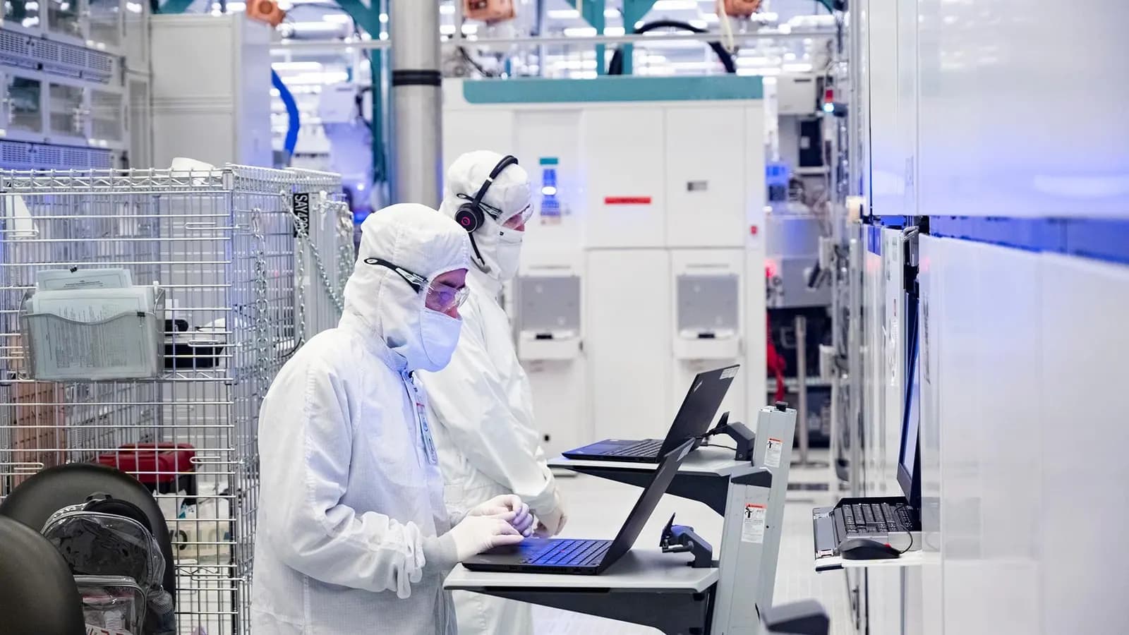 Two workers in white coveralls and masks monitor semiconductor manufacturing equipment on laptops in a cleanroom.