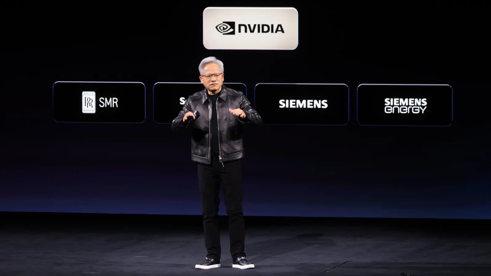 Sahnede siyah deri ceketli bir konuşmacı; arkadaki ekranda NVIDIA, Rolls-Royce SMR, Siemens ve Siemens Energy logoları görünüyor.