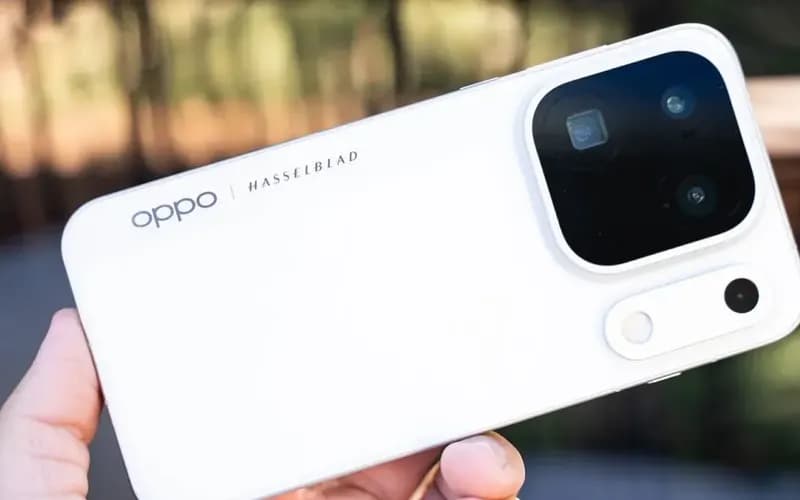 Beyaz bir Oppo telefonun Hasselblad yazılı arka yüzü ve kare kamera modülü, elde tutulurken bulanık doğa arka planında görünüyor