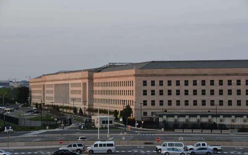 Alacakaranlıkta büyük, bej taş cepheli resmi bir bina; önünde otopark, güvenlik kulübesi ve park halindeki araçlar görülüyor.