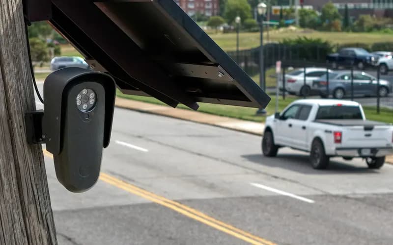 Solar-powered security camera mounted on a pole, with a white pickup truck driving down the street in the background