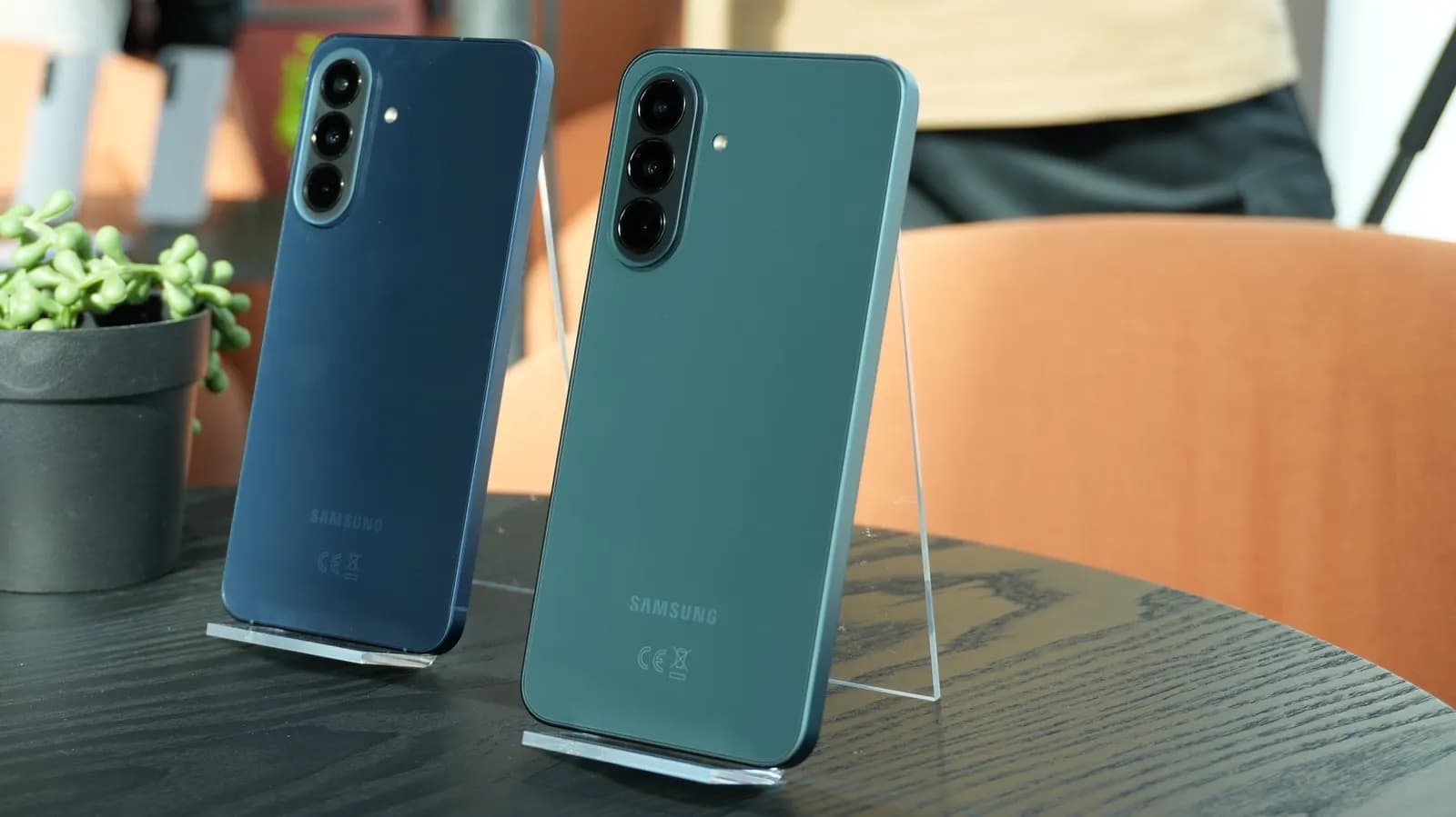 Siyah masada akrilik altlıklar üzerinde duran, mavi ve yeşil renkli iki Samsung akıllı telefonun arka yüzü
