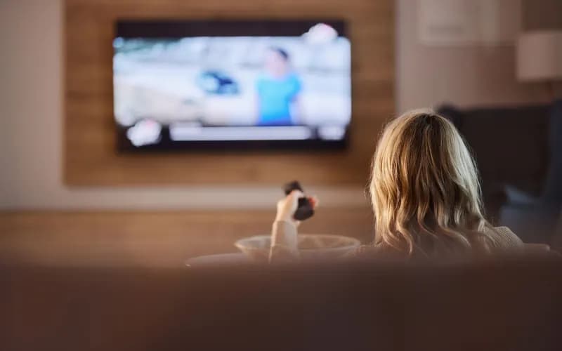 Sarı saçlı bir kadın koltukta oturup elindeki kumandayla duvardaki televizyonu izliyor; ekran bulanık.