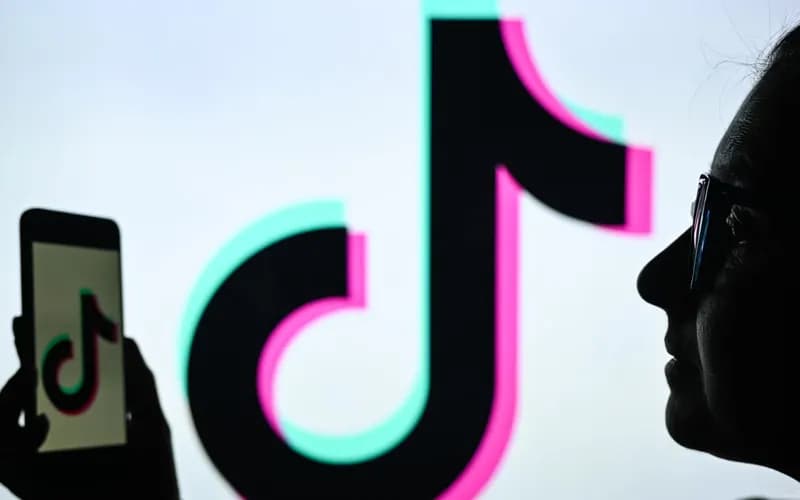 Gözlüklü bir kadının silueti, elindeki telefonda ve arkadaki dev ekranda TikTok logosu görünüyor.