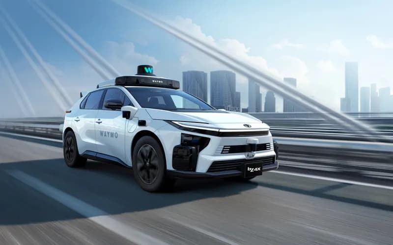 Waymo logolu beyaz otonom Toyota bZ4X, arkada gökdelenler bulunan bir yolda hızla ilerliyor.