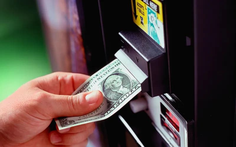 Bir el, otomat makinesinin banknot okuyucusuna bir dolarlık banknotu sokuyor.